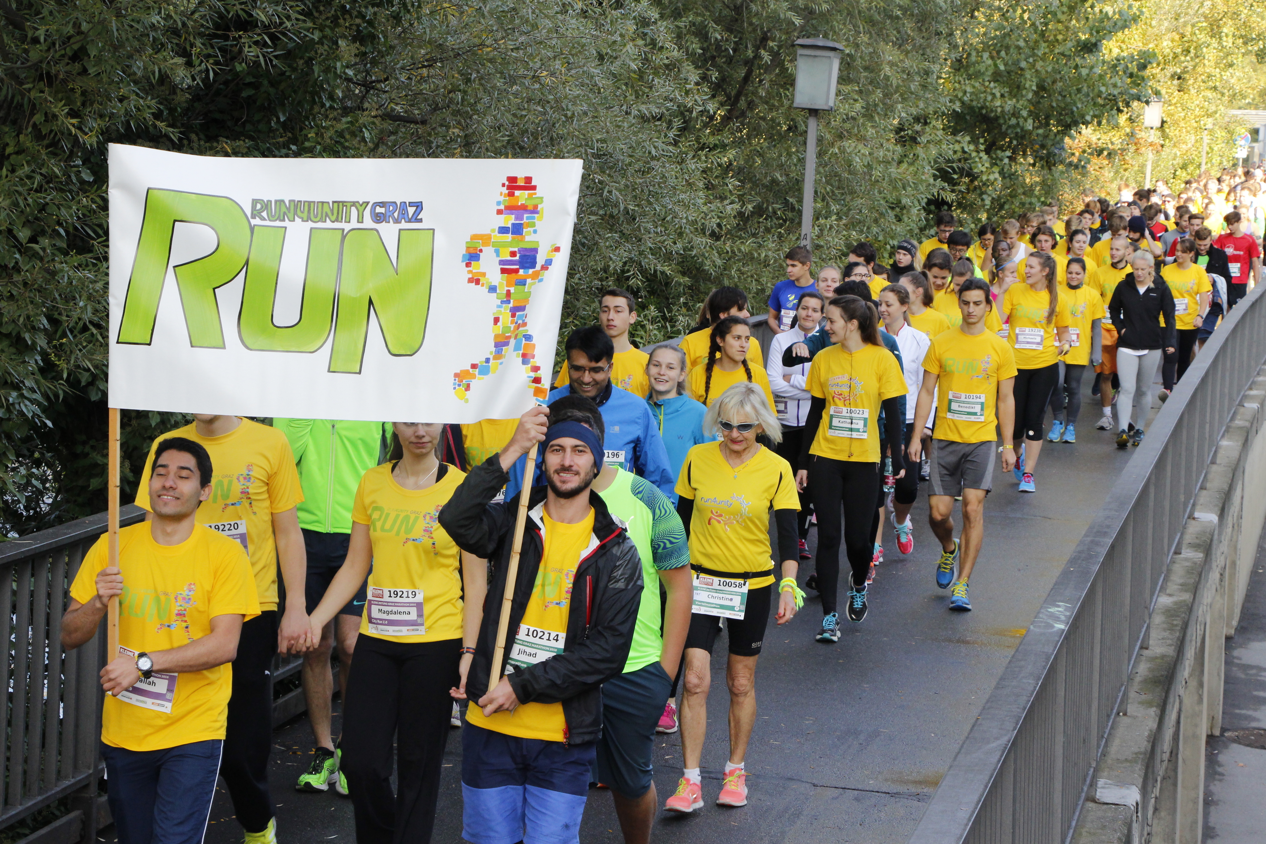 Läufer: innen des Run4unity mit gelben T-Shirts und Plakat im Vordergrund mit Veranstaltungslogo, das von Läufer: innen mit getragen wird.